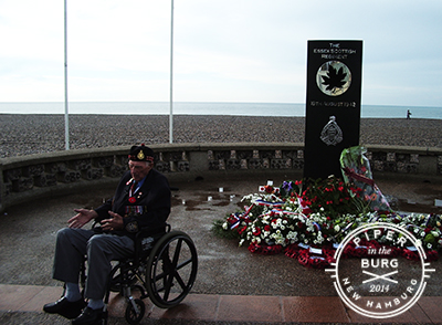 Veteran wearing glengarry and jacket sits in wheelchair next to black modern monument on the rocky shoreline