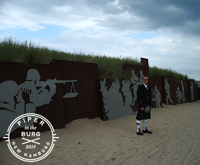 Bagpiper standing on path to beach in front of metal wall depicting solider firing a gun