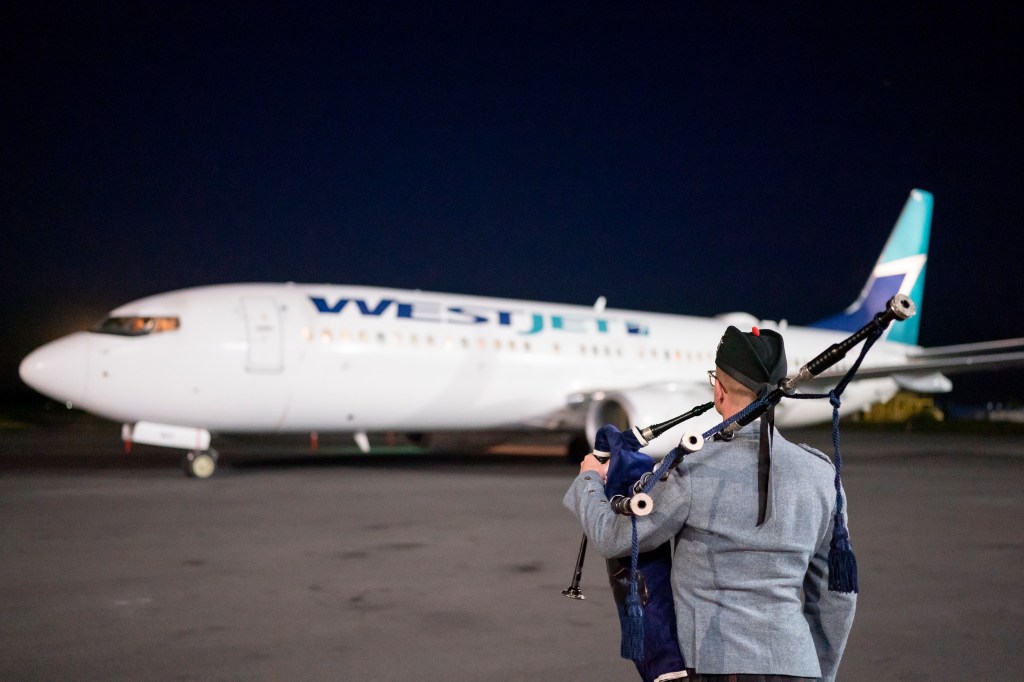 Andrew Osborne - Halifax Bagpiper standing facing a WestJet airplane. 